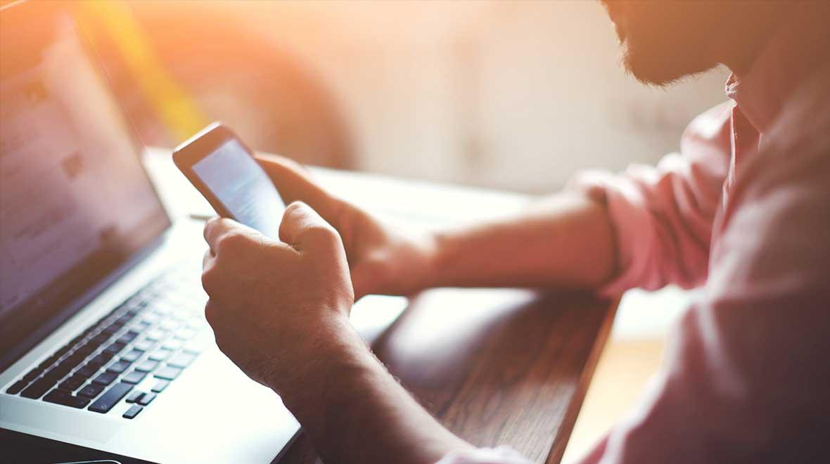 Pessoa usando smartphone com laptop ao fundo