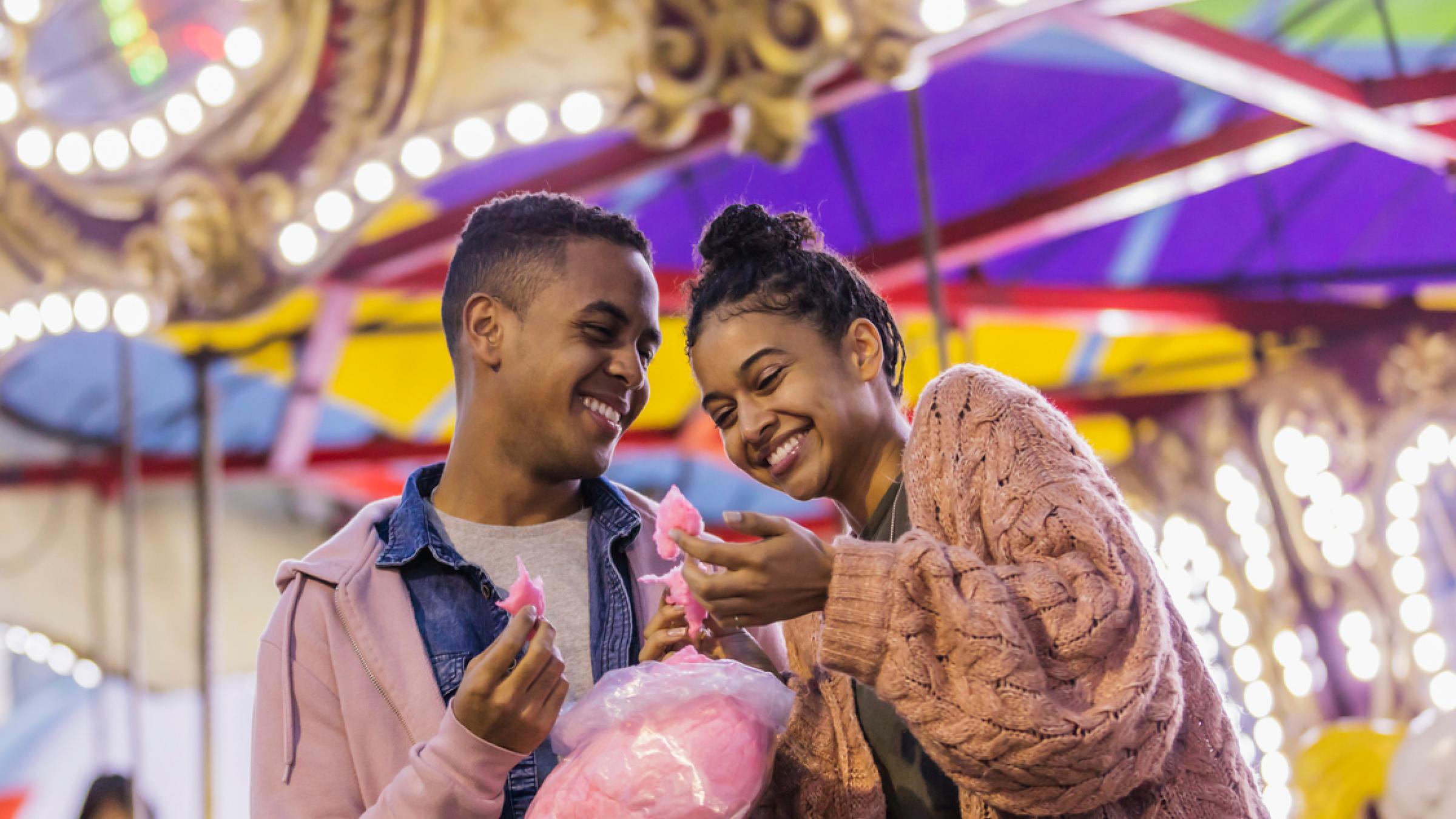 Two people sharing cotton candy and smiling at each other at a carnival.