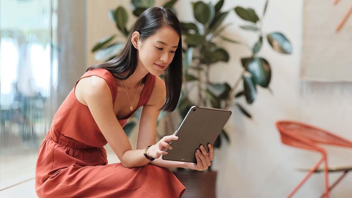 Woman in a red dress sitting and using a tablet in a modern space with plants in the background.