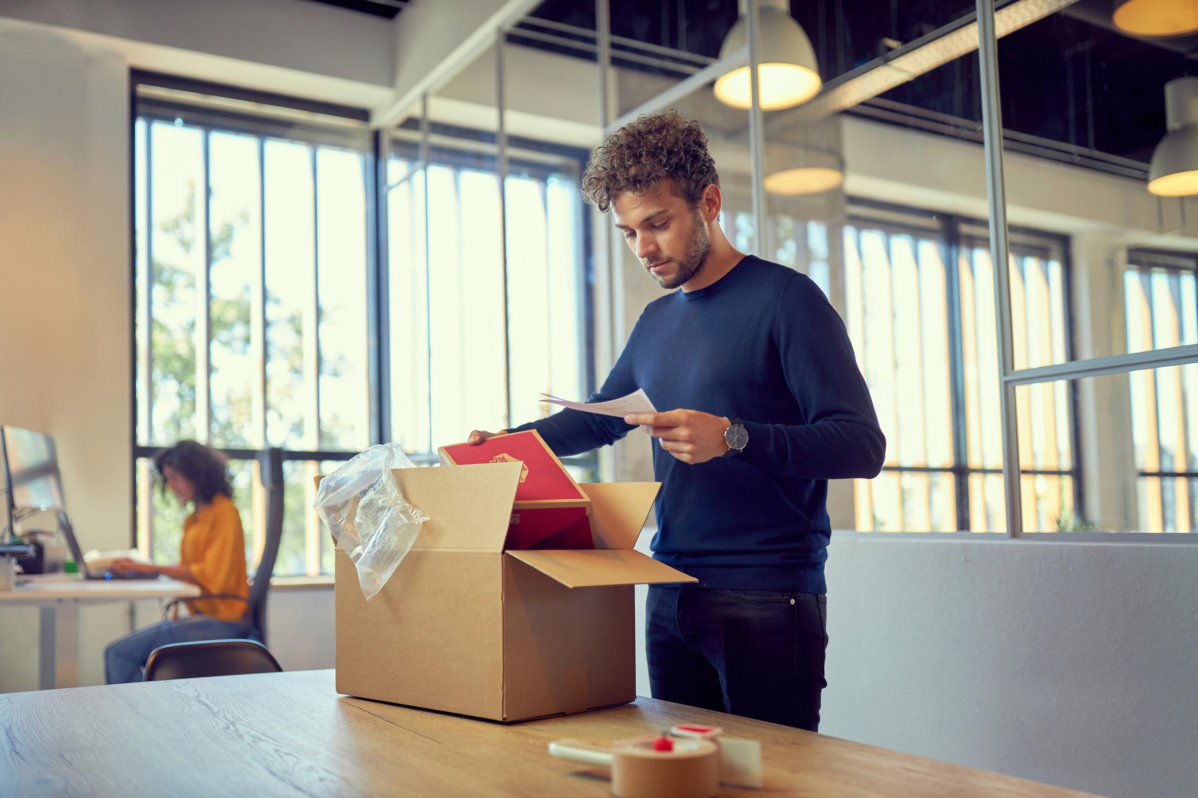 A man unpacking a box, holds a smaller box while looking to a paper in his hand.