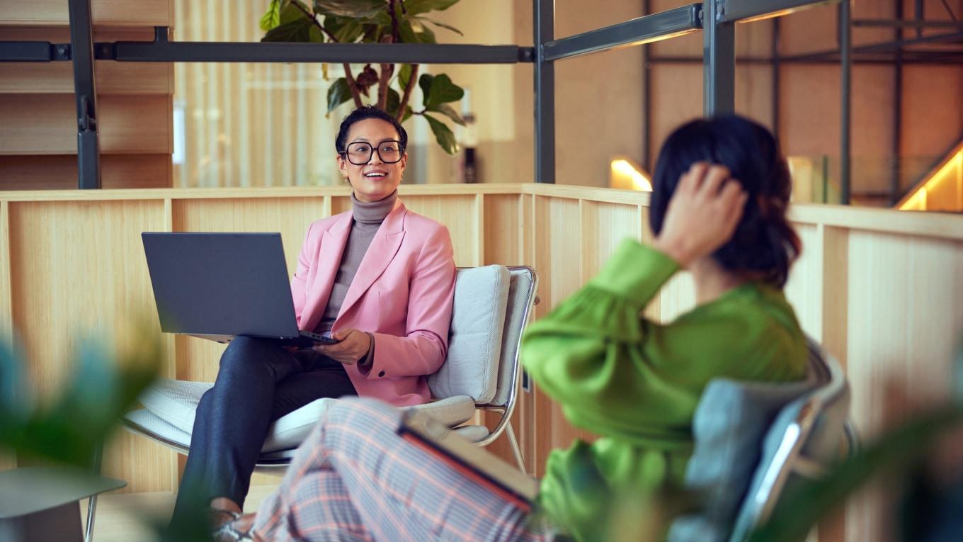 Two women in a modern office setting, one working on a laptop and the other in a conversation.