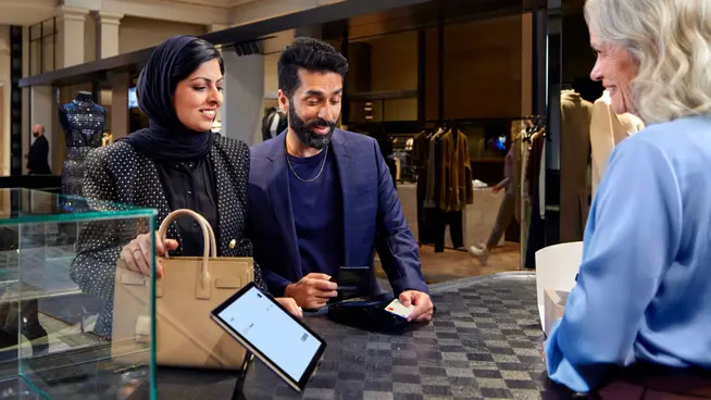 Two customers interacting with a salesperson using an Adyen payment terminal on a tablet at a retail store checkout.