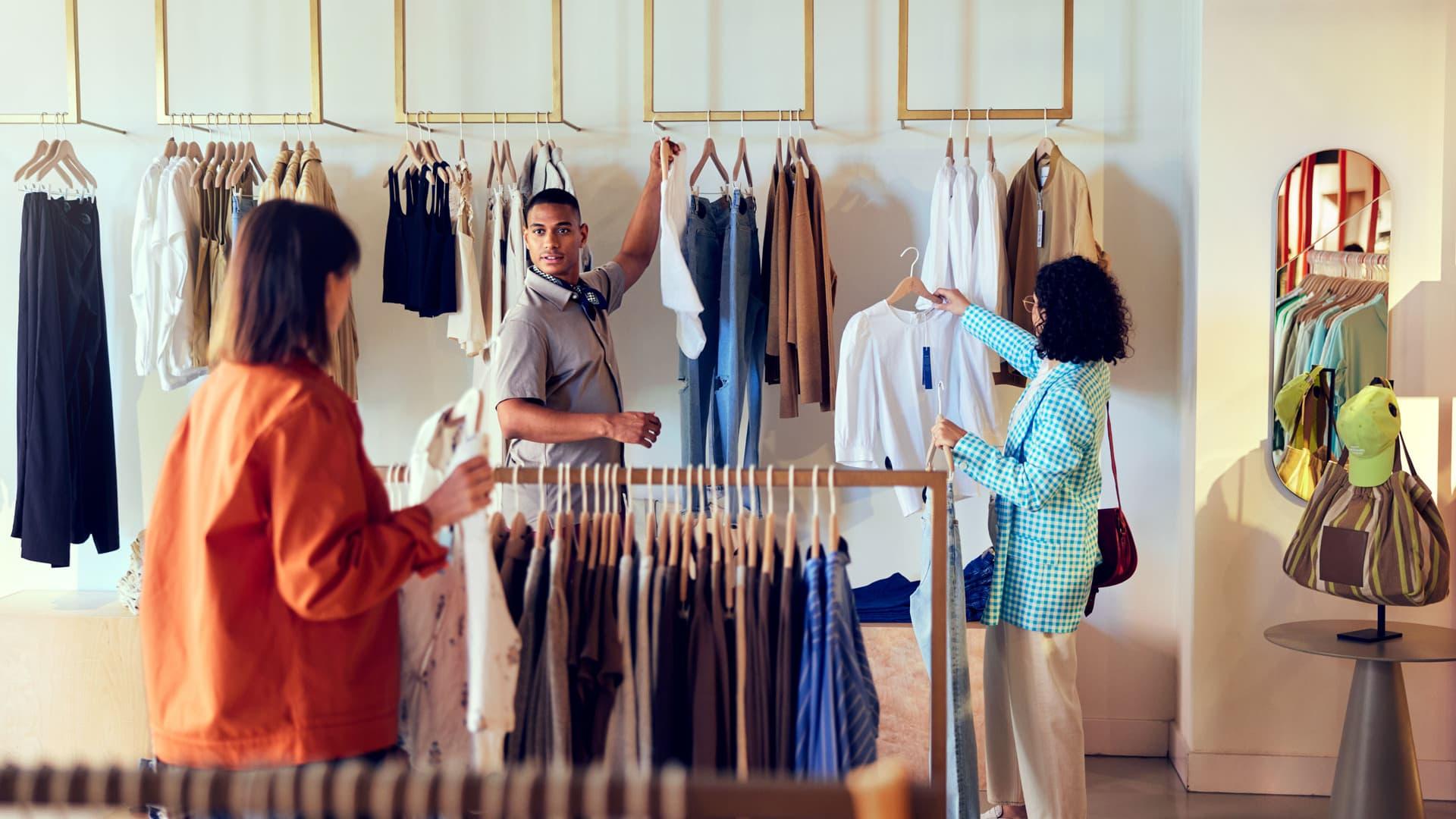 Several clothes hangers hanging with a salesman holding them in the background and two women watching