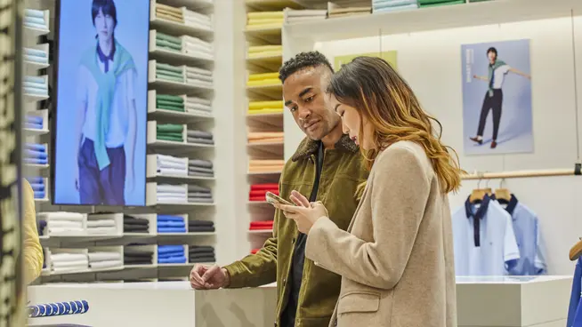 Two people looking at a smartphone in a clothing store with shelves of shirts in the background.