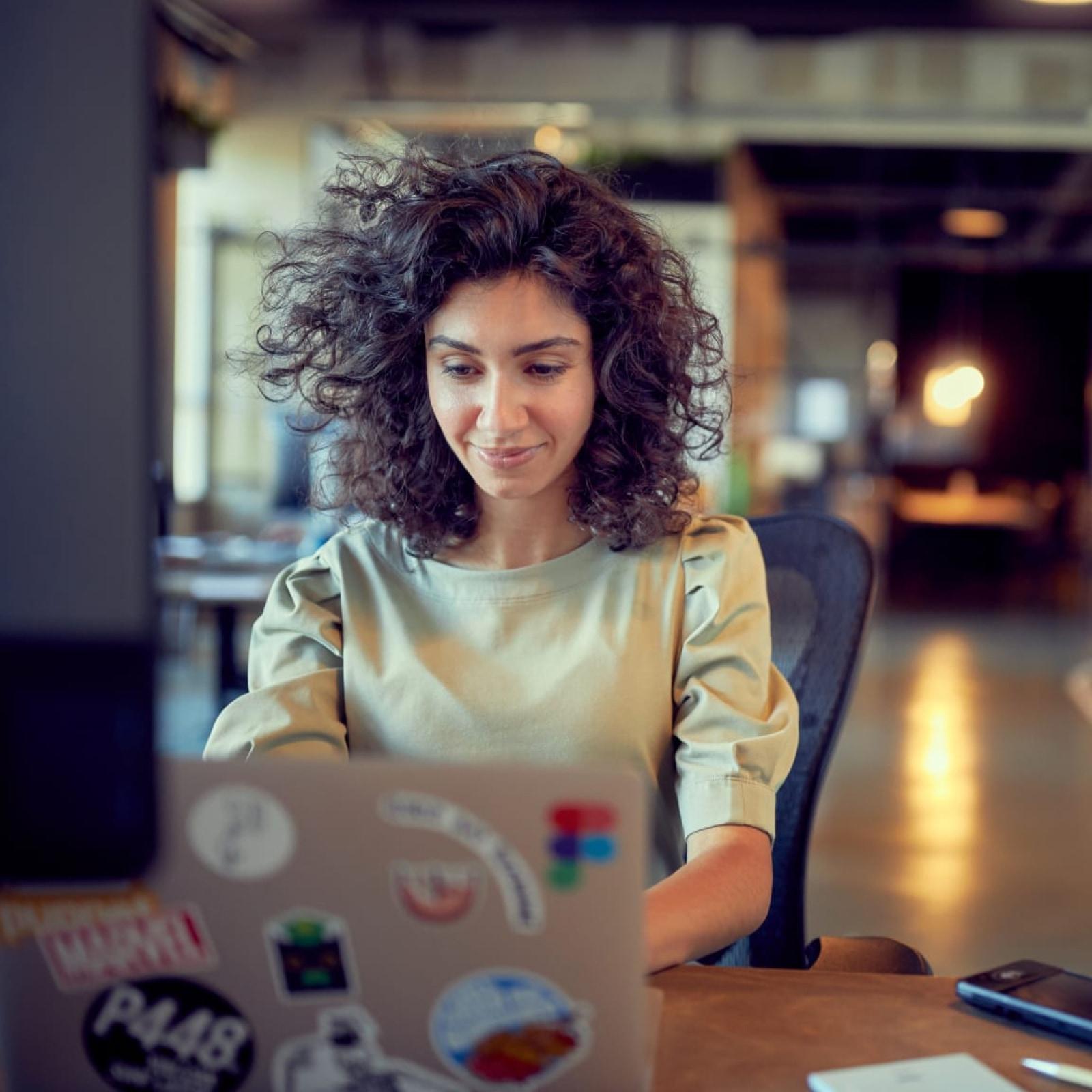 Mulher sorrindo trabalhando em um laptop com adesivos, possivelmente em um ambiente de escritório informal.