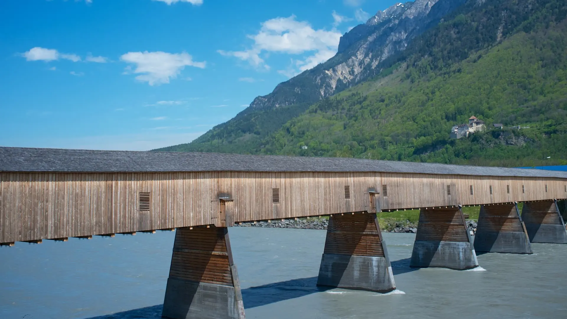 A long wooden covered bridge with concrete pillars over a river, mountains and a castle in the background in Liechtenstein.