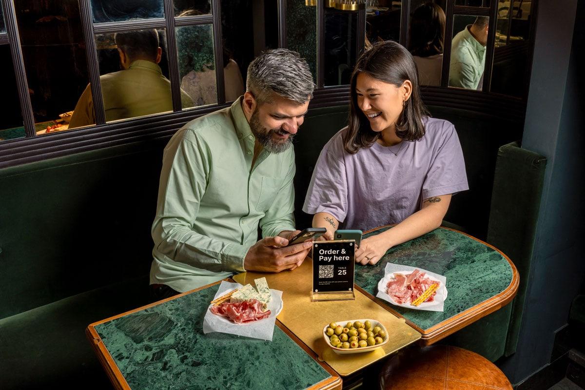 Two people at a restaurant table using a smartphone with a tabletop sign for ordering and payment.
