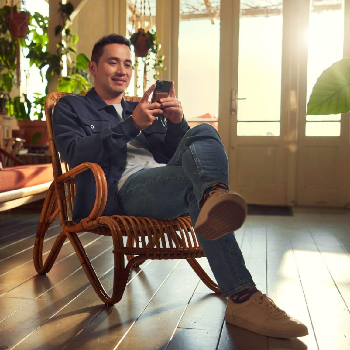 Homem sentado em uma cadeira de rattan usando um smartphone em um quarto com plantas e luz solar.