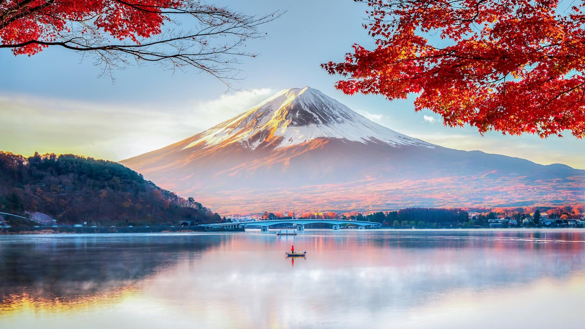 Mount Fuji in Japan with snow cap reflected in a lake with red autumn leaves in the foreground.