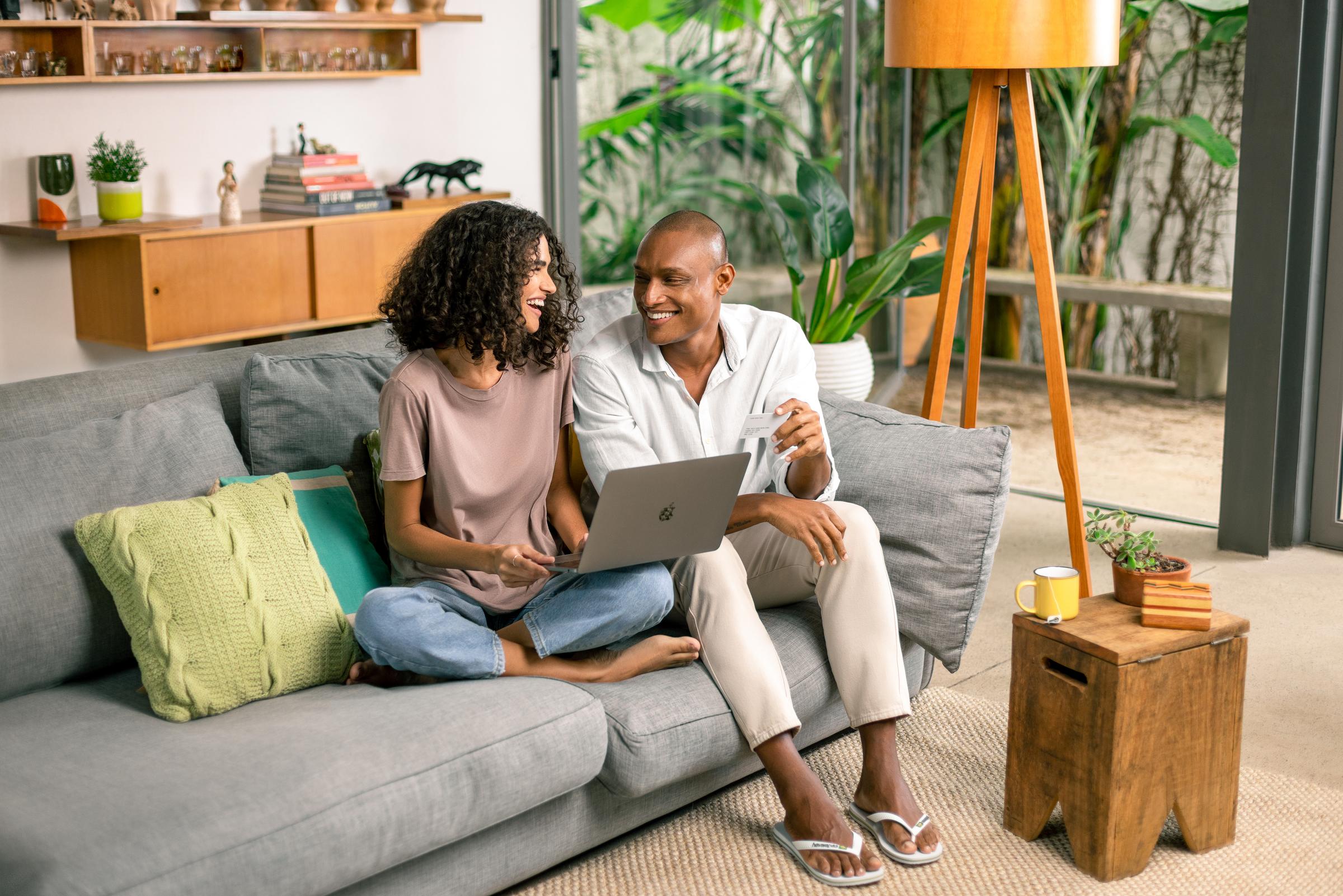 Um homem e uma mulher sentados no sofá, segurando um laptop e um cartão de crédito.
