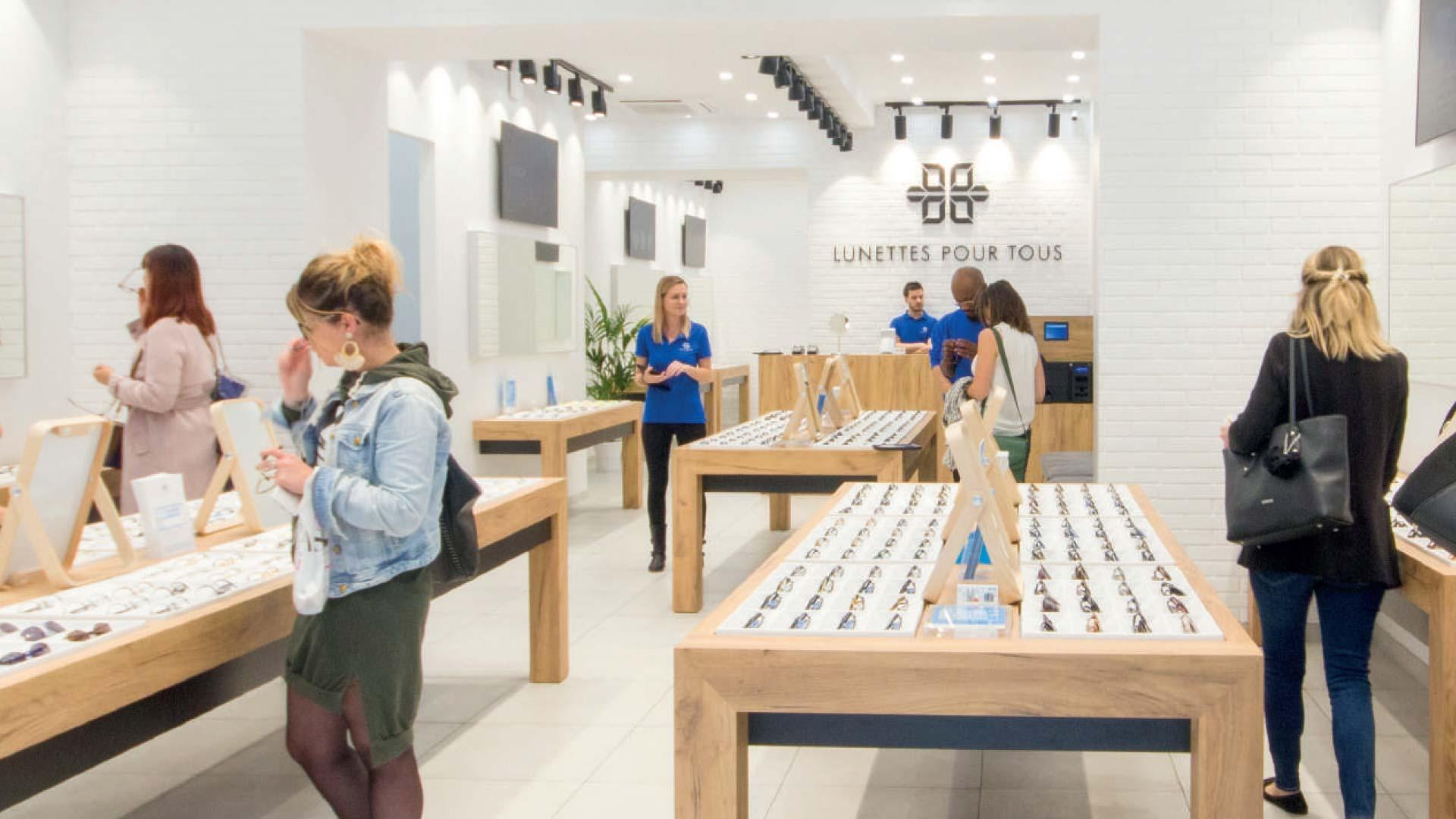 Clients et personnel dans un magasin d'optique moderne avec des vitrines de lunettes sur des tables en bois.