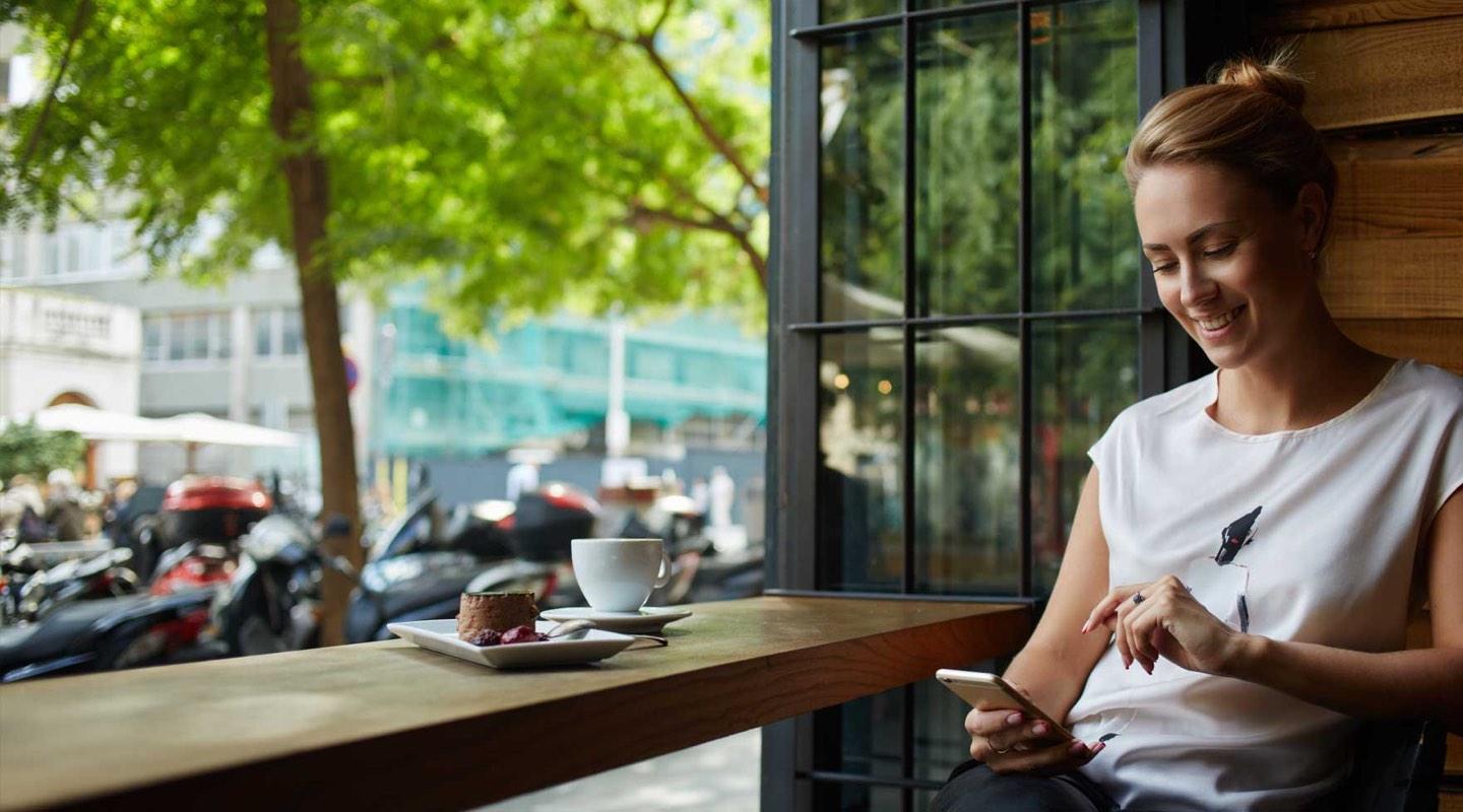 Femme souriante assise à une table de café utilisant un smartphone avec café et gâteau.