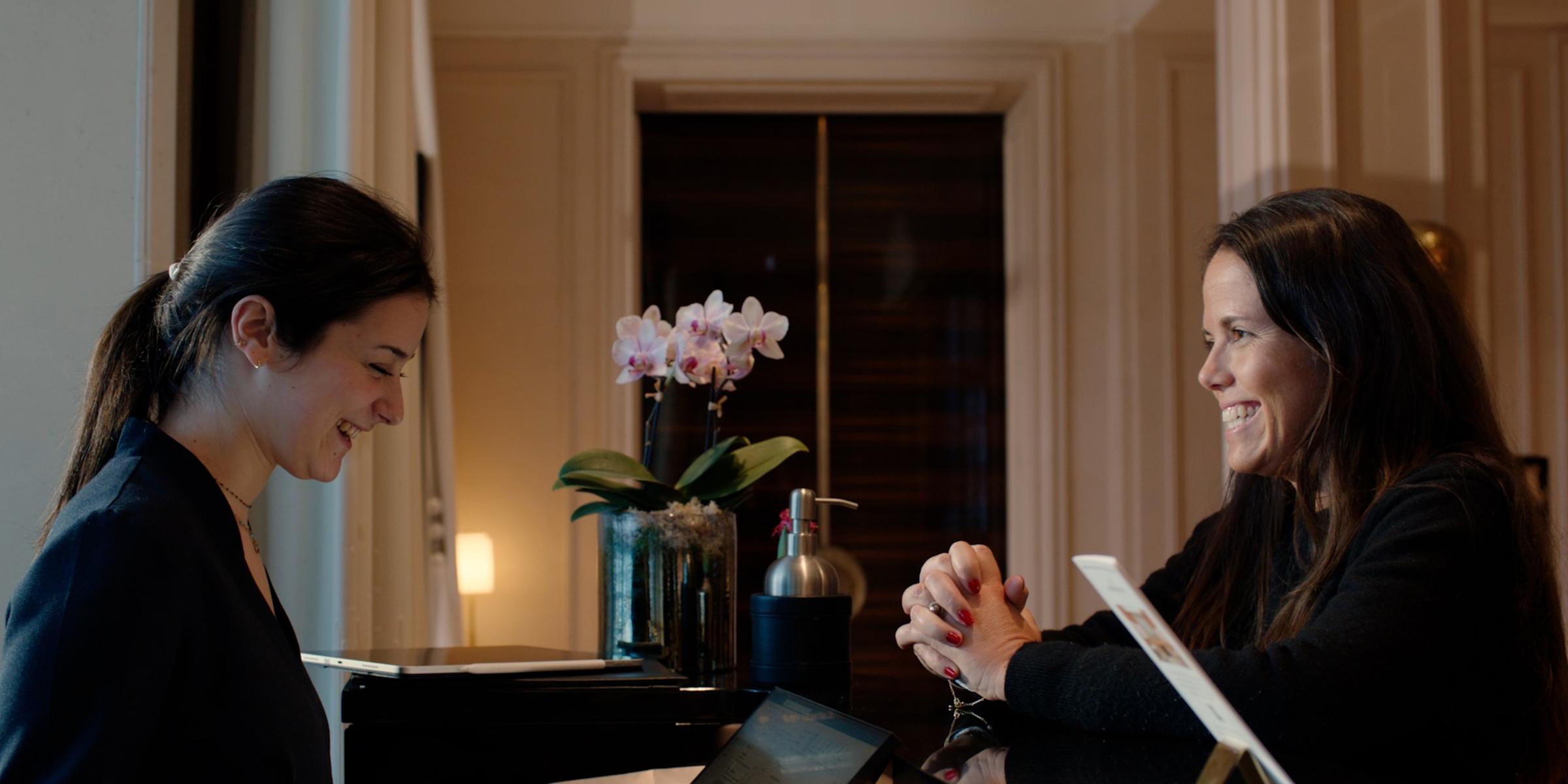 Two women smiling at a reception desk with one holding a payment terminal.