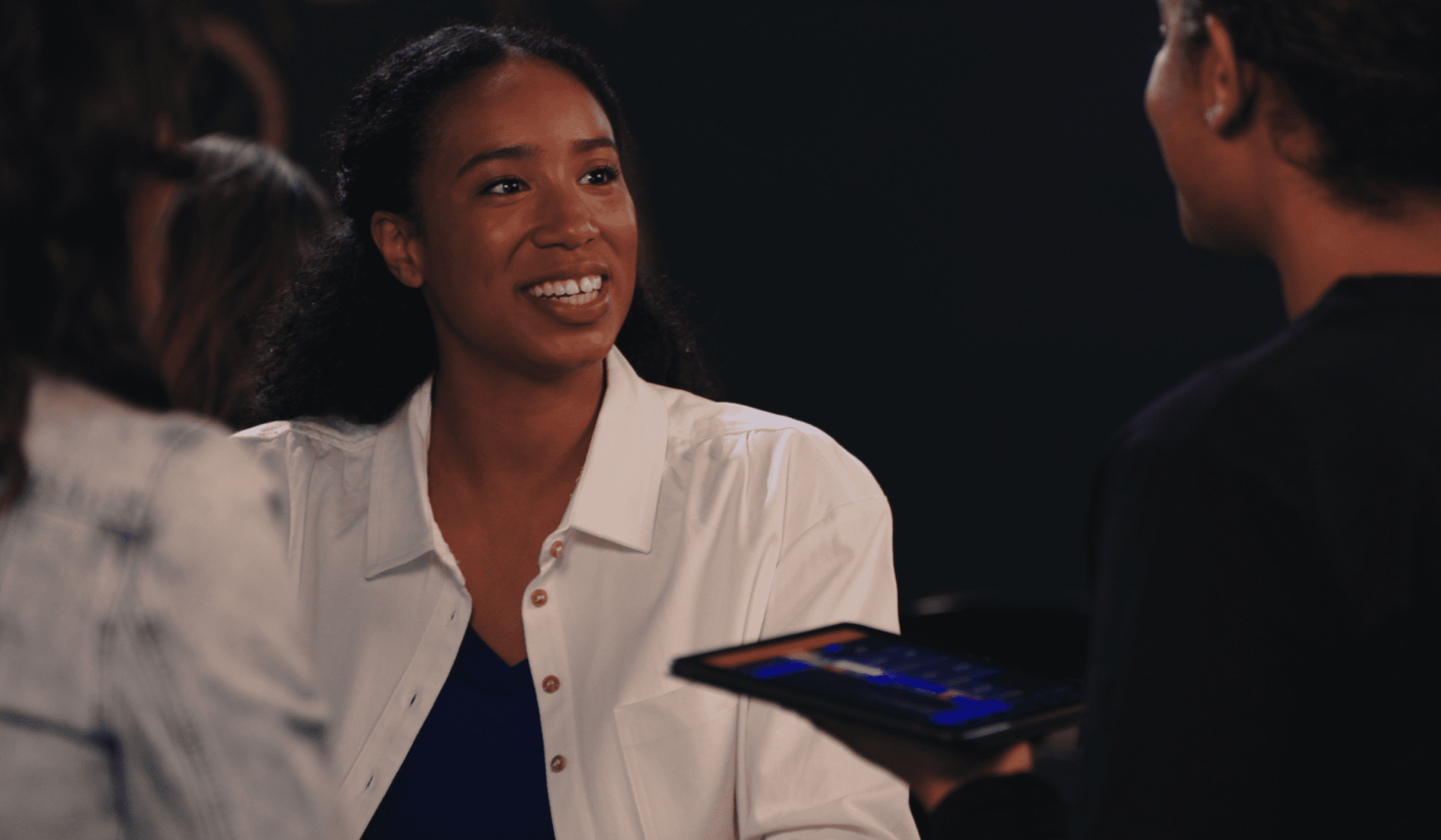 Smiling woman engaged in conversation with colleagues using a tablet displaying a payment application.