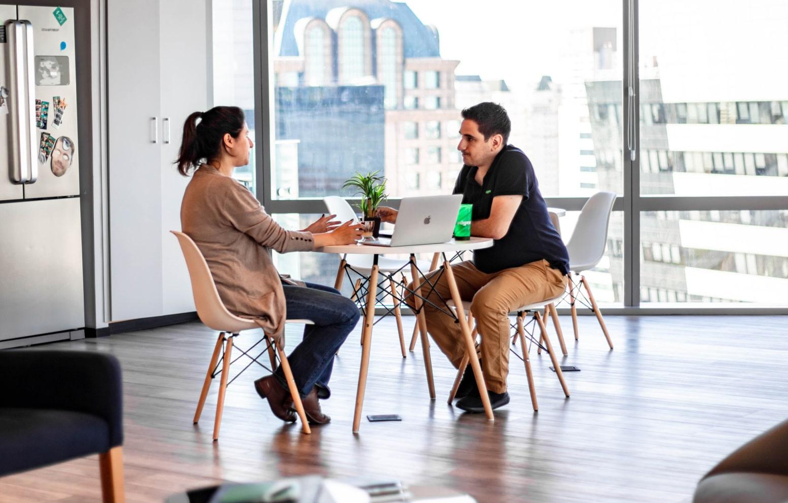 Dos personas sentadas en una mesa con portátiles en una oficina moderna con paisaje urbano de fondo.