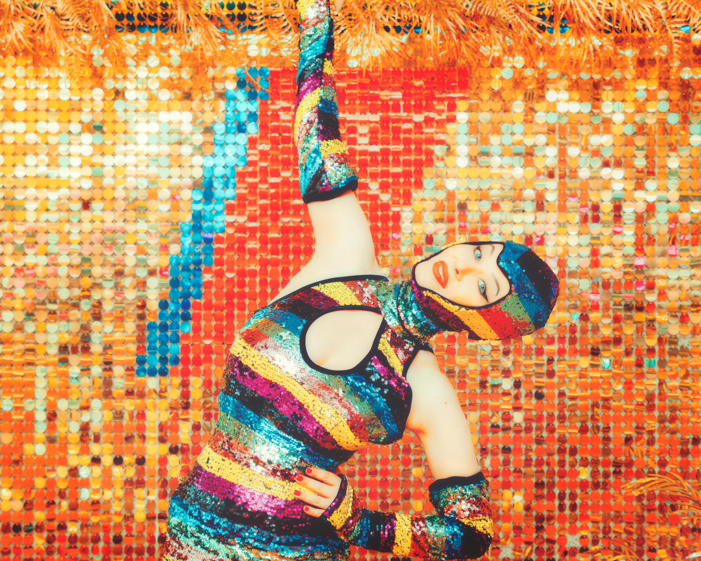 A dancer wearing a rainbow glitter outfit on a bright orange background