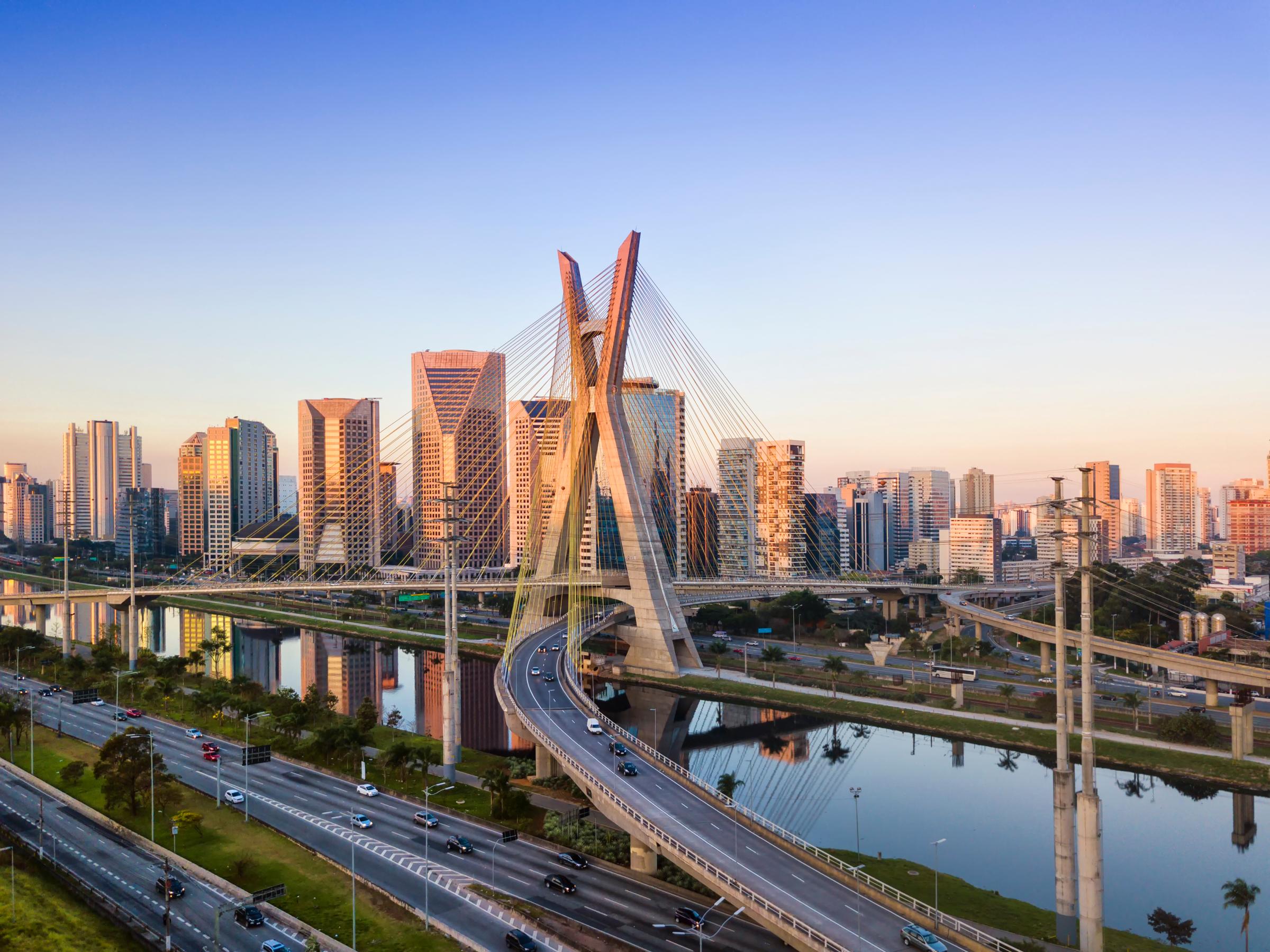 A cityscape with modern buildings and a cable-stayed bridge over a river at sunset