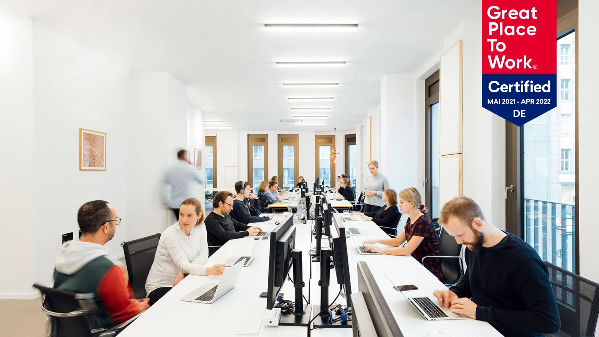Mitarbeiter arbeiten in einem hellen Büroraum mit einem Great Place to Work-zertifizierten Banner