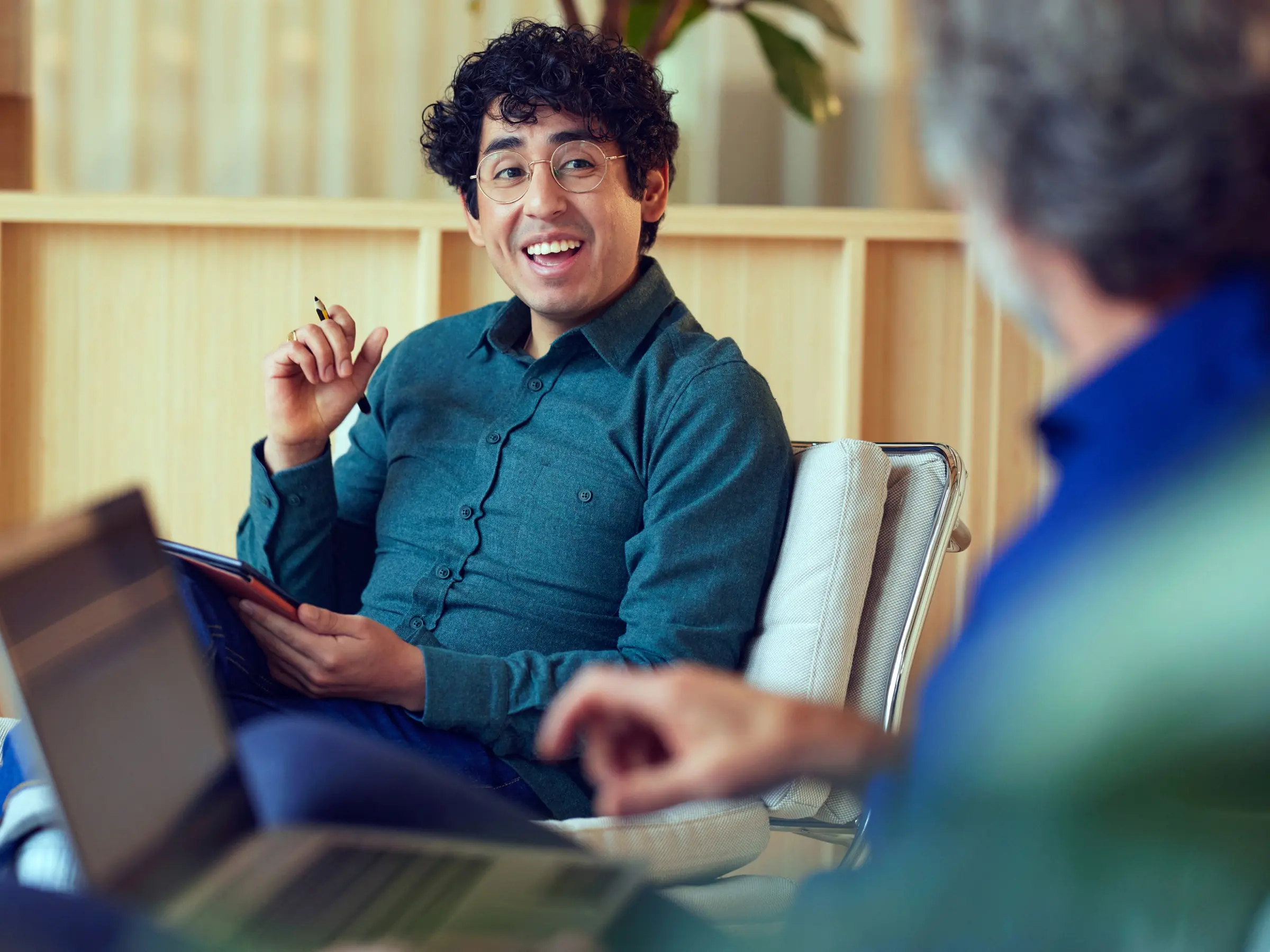 Smiling person holding a pen and digital tablet during a meeting with another individual.