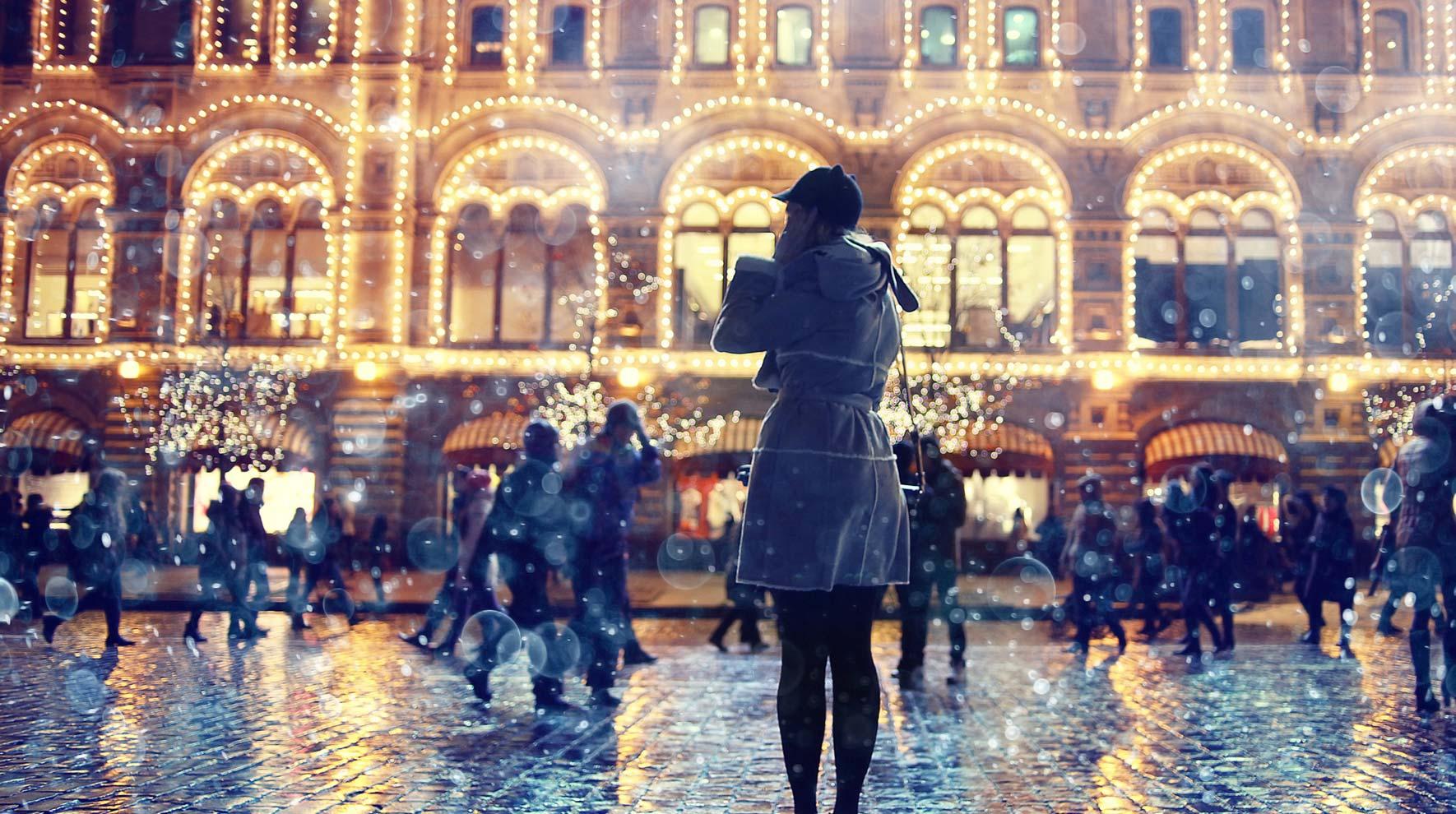 Personne debout devant un bâtiment illuminé avec des lumières festives et une foule occupée au crépuscule.