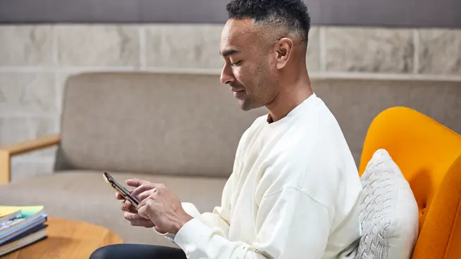 Man sitting on a couch using a smartphone with a book and orange chair nearby.