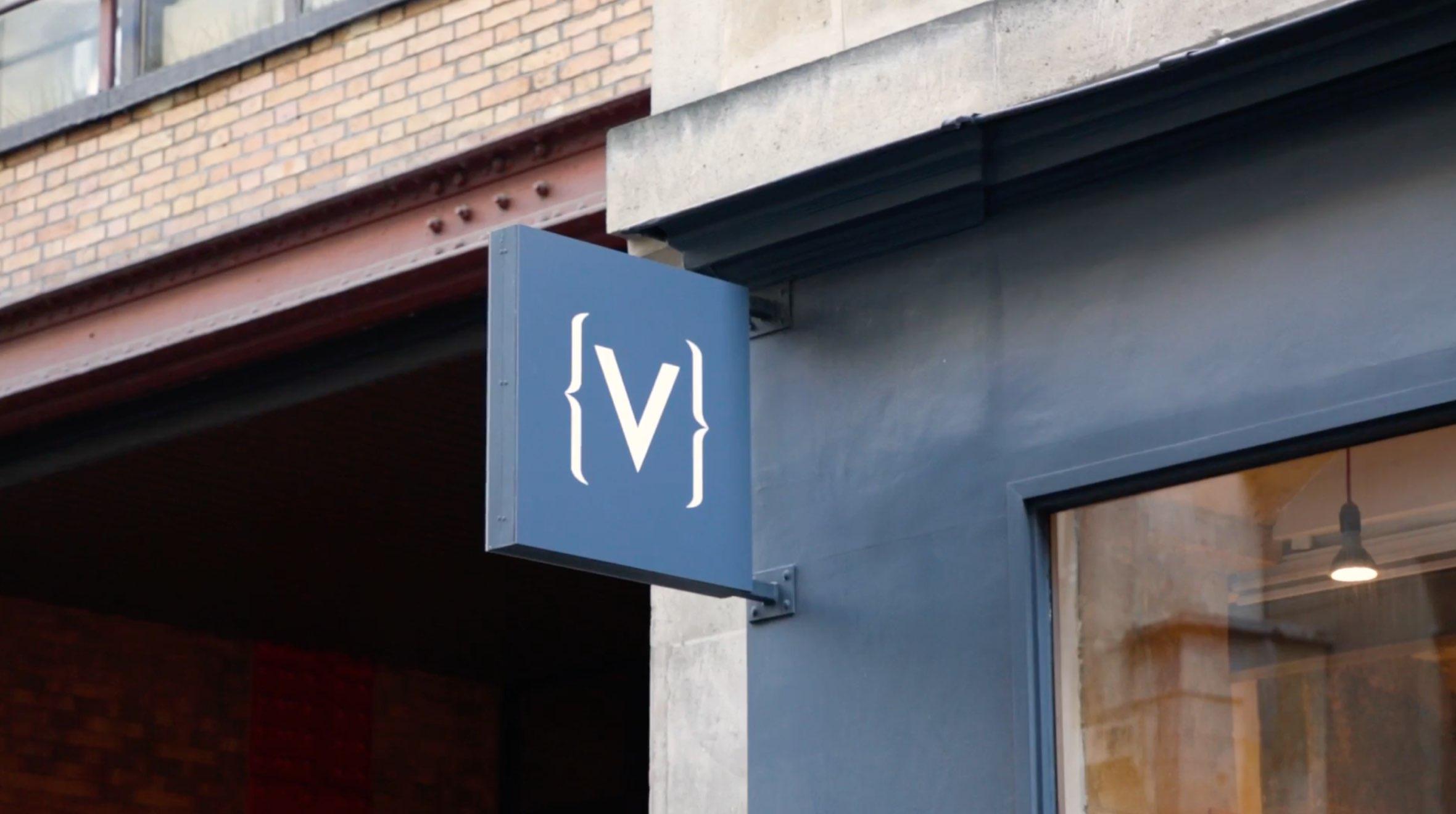 Panneau carré avec lettre 'V' stylisée suspendue à l'extérieur d'un bâtiment.