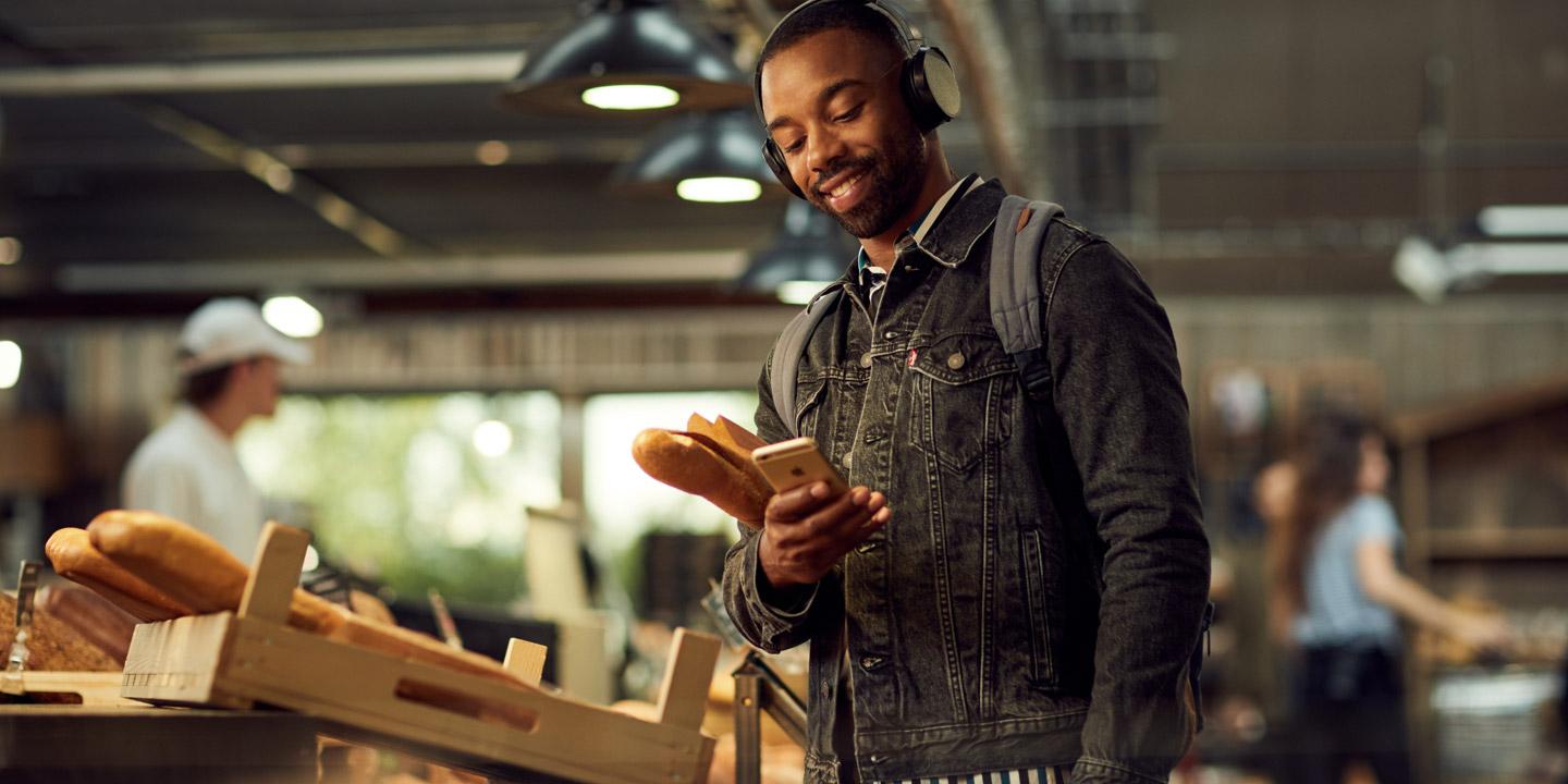 Man ler mot telefonen med en baguette i en bageri med anställda i bakgrunden.