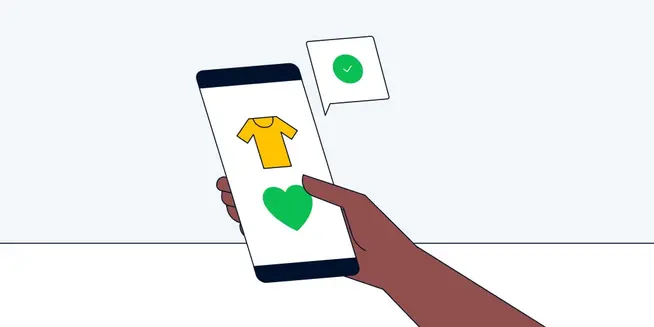 Mano sosteniendo un smartphone que muestra un diseño de camiseta y un icono de corazón verde con una burbuja de verificación.
