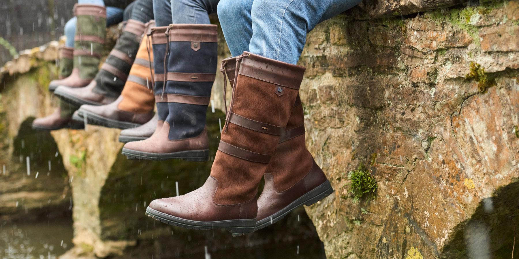 Three pairs of legs dangling over a wall wearing various styles of boots in the rain.