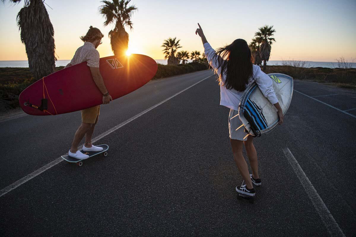 Zwei Personen mit Surfbrettern auf einer Küstenstraße bei Sonnenuntergang.