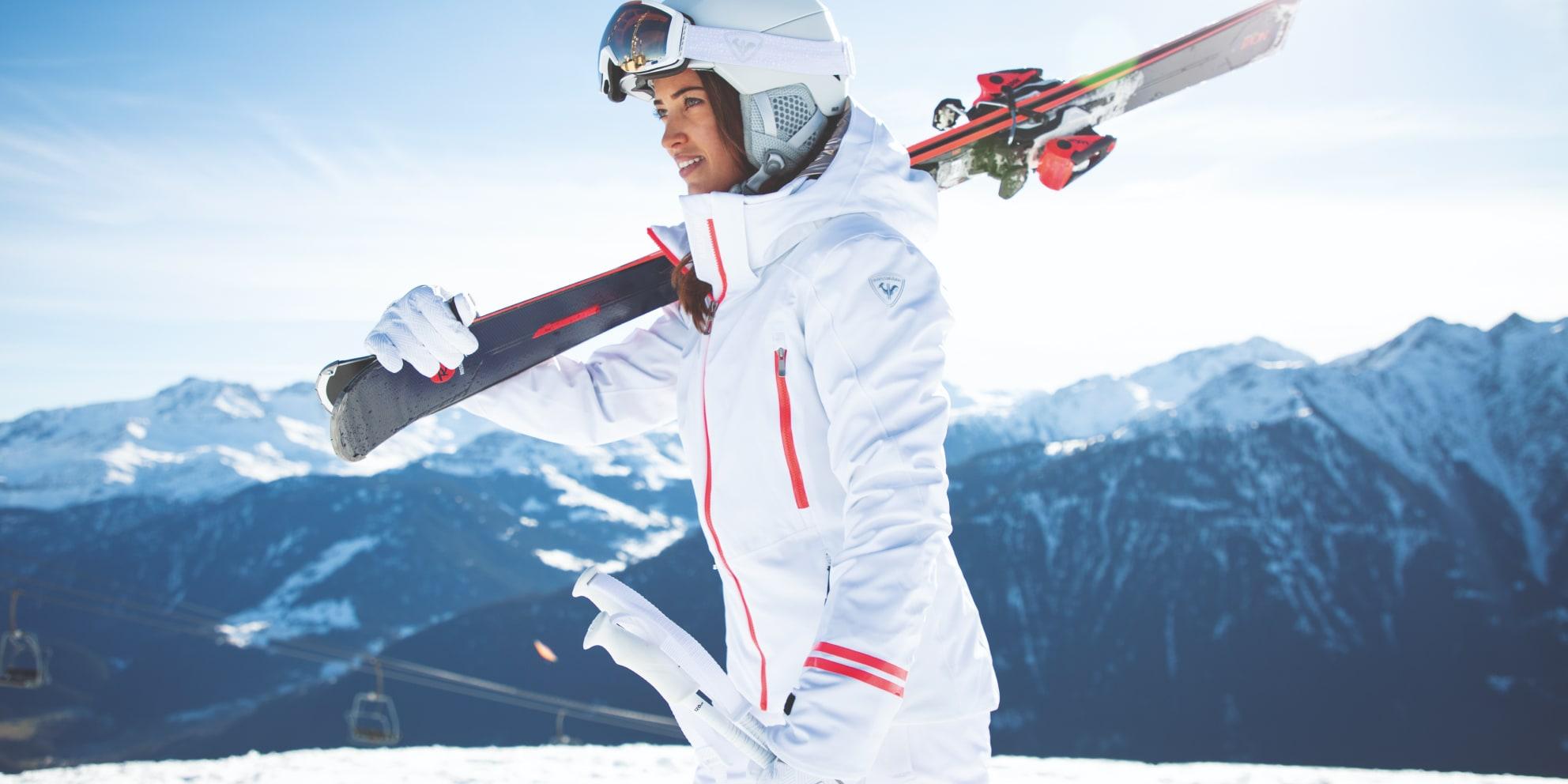 Skieuse portant des skis sur son épaule avec des montagnes enneigées en arrière-plan.