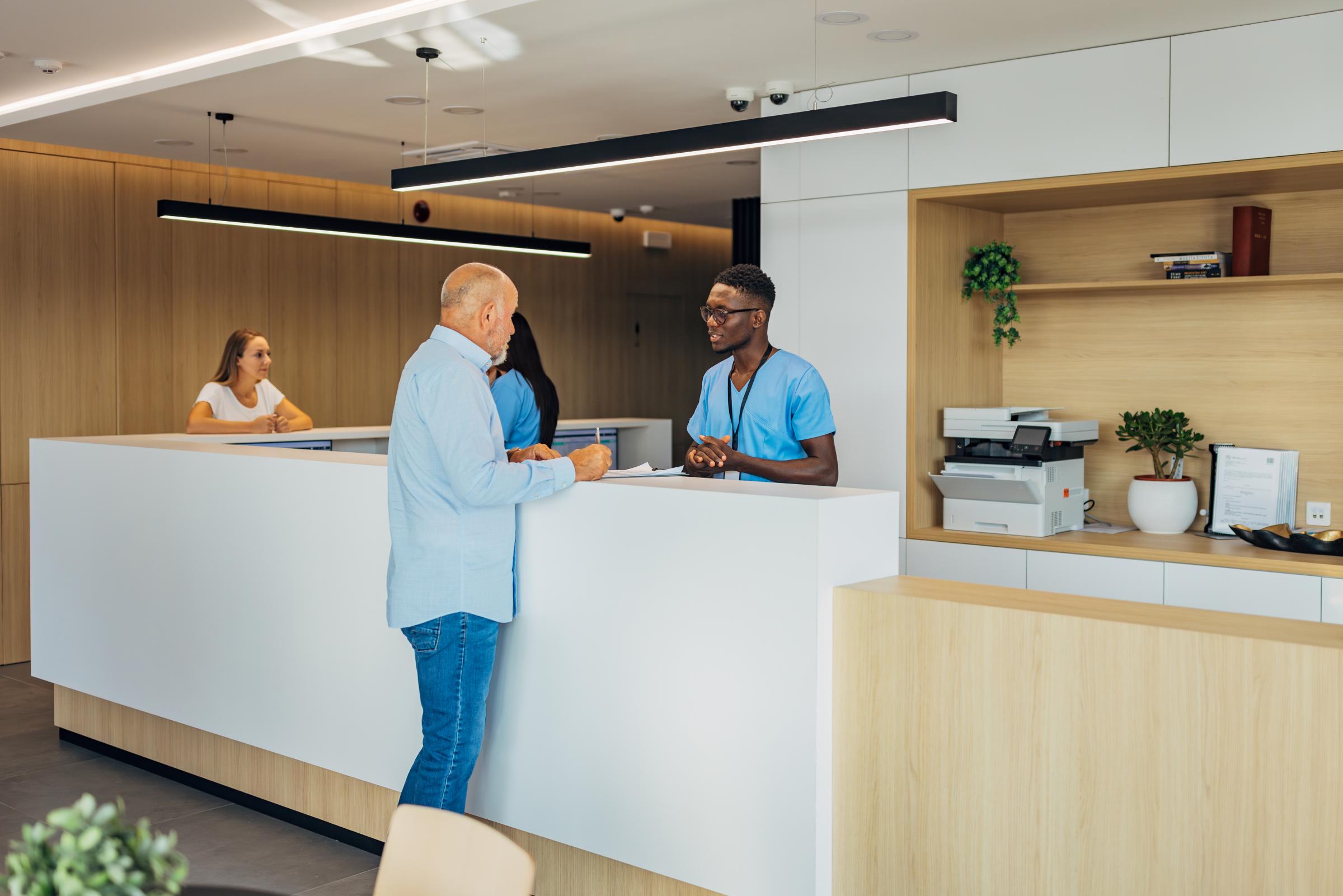 Medical worker at the clinic reception explains to a senior male patient how to fill out a form before starting his hospital treatment