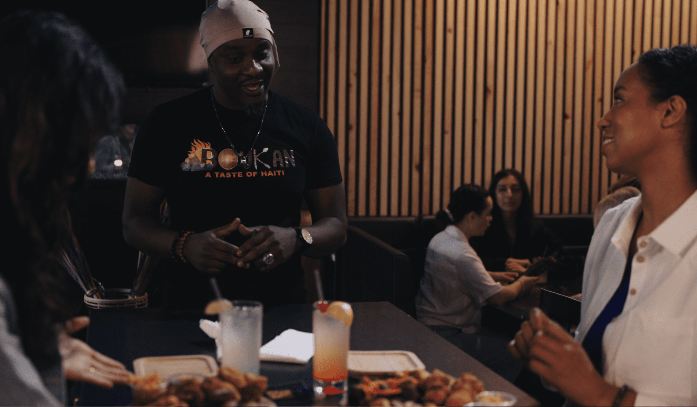 Waiter wearing a 'BOUKAN A Taste of Haiti' t-shirt talking to customers in a restaurant.