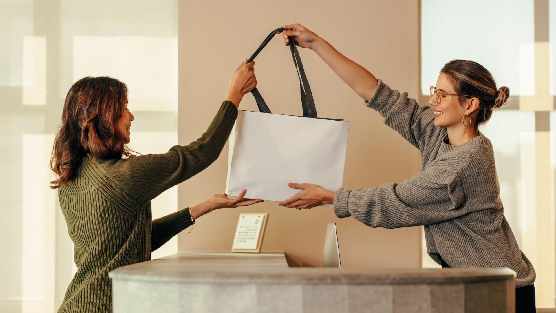 Two people smiling at each other while exchanging a white shopping bag over a counter.