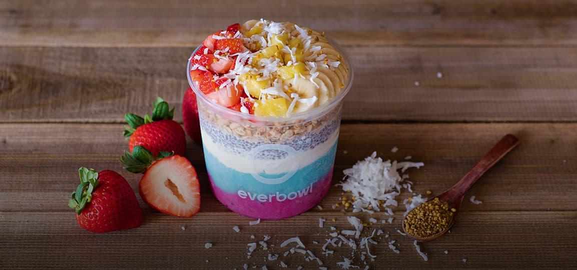 Bol de frutas en capas con fresas y coco rallado, al lado de frutas frescas y una cuchara de madera con semillas.