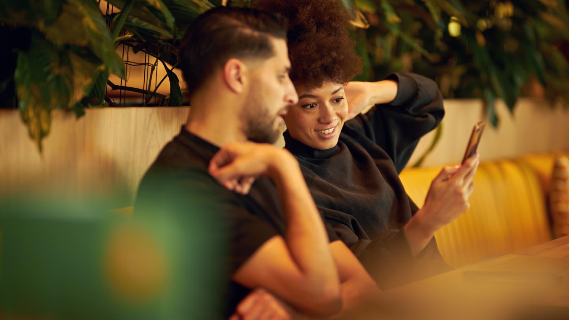 Two people cozying up on a couch with one showing something on a mobile phone to the other.