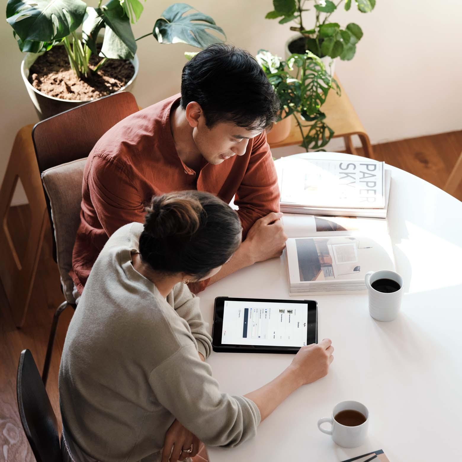 Two people examining a tablet at a white table with coffee and reading materials.