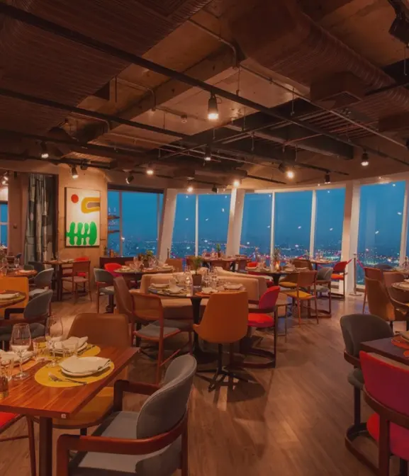 Interior elegante de restaurante ao entardecer com mesas postas para jantar e luzes da cidade ao fundo.