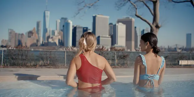 Zwei Frauen entspannen sich in einem Außen-Infinitypool mit Stadtskyline im Hintergrund.