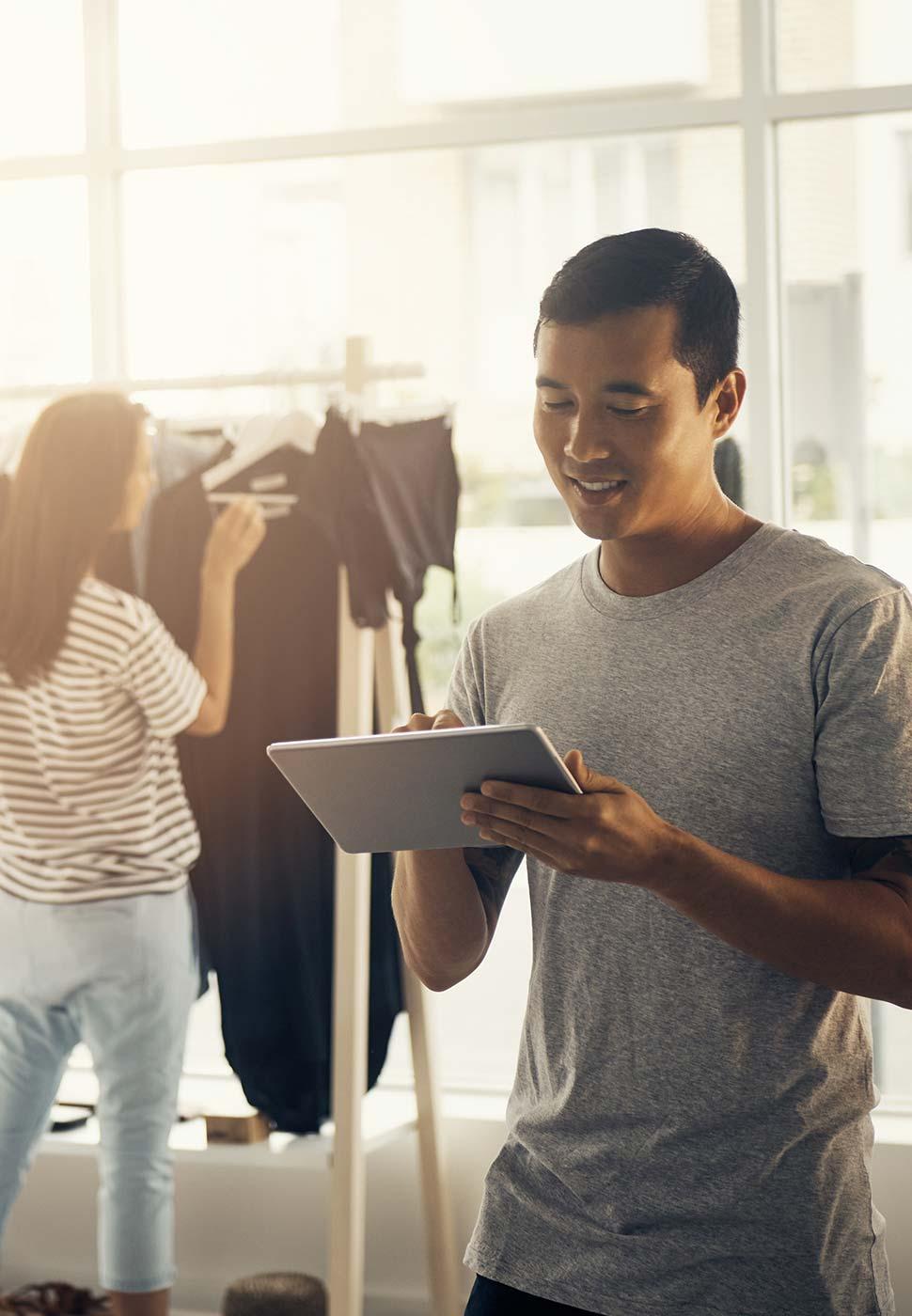 Hombre utilizando una tableta en una tienda de ropa brillante con una mujer revisando ropa en el fondo.