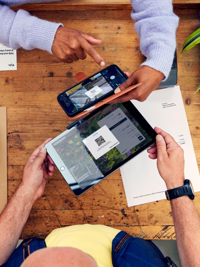 Duas pessoas a usar smartphones para digitalizar um código QR no ecrã de um tablet acima de documentos em cima de uma mesa de madeira.