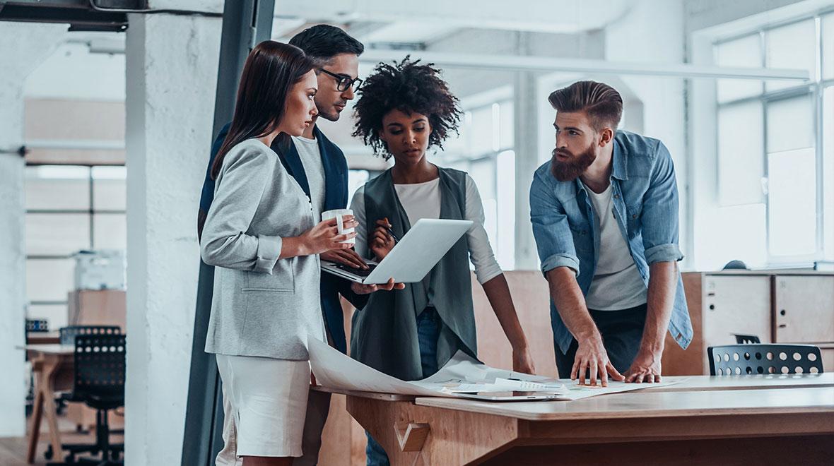 Quatro profissionais engajados em uma discussão com laptop e papéis em um escritório moderno.