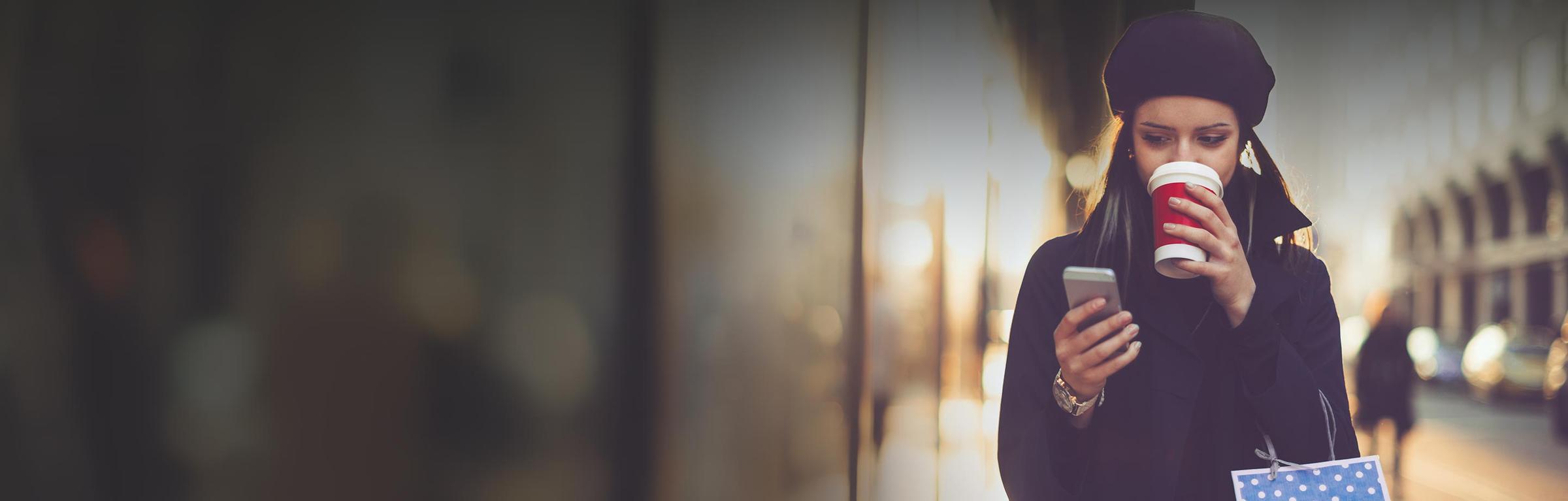 Woman in a beret drinking a coffee and looking at her phone on a city street.