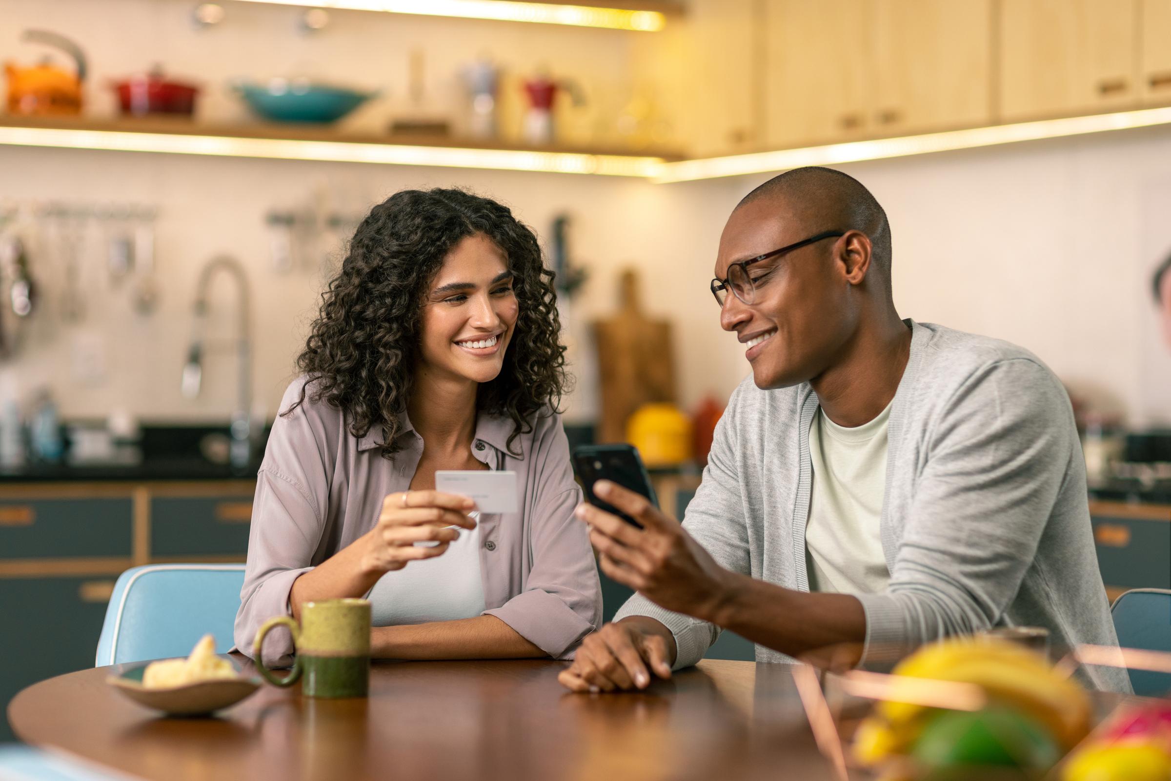 Homem e mulher sentados à mesa da cozinha segurando um cartão de crédito e um telefone celular.