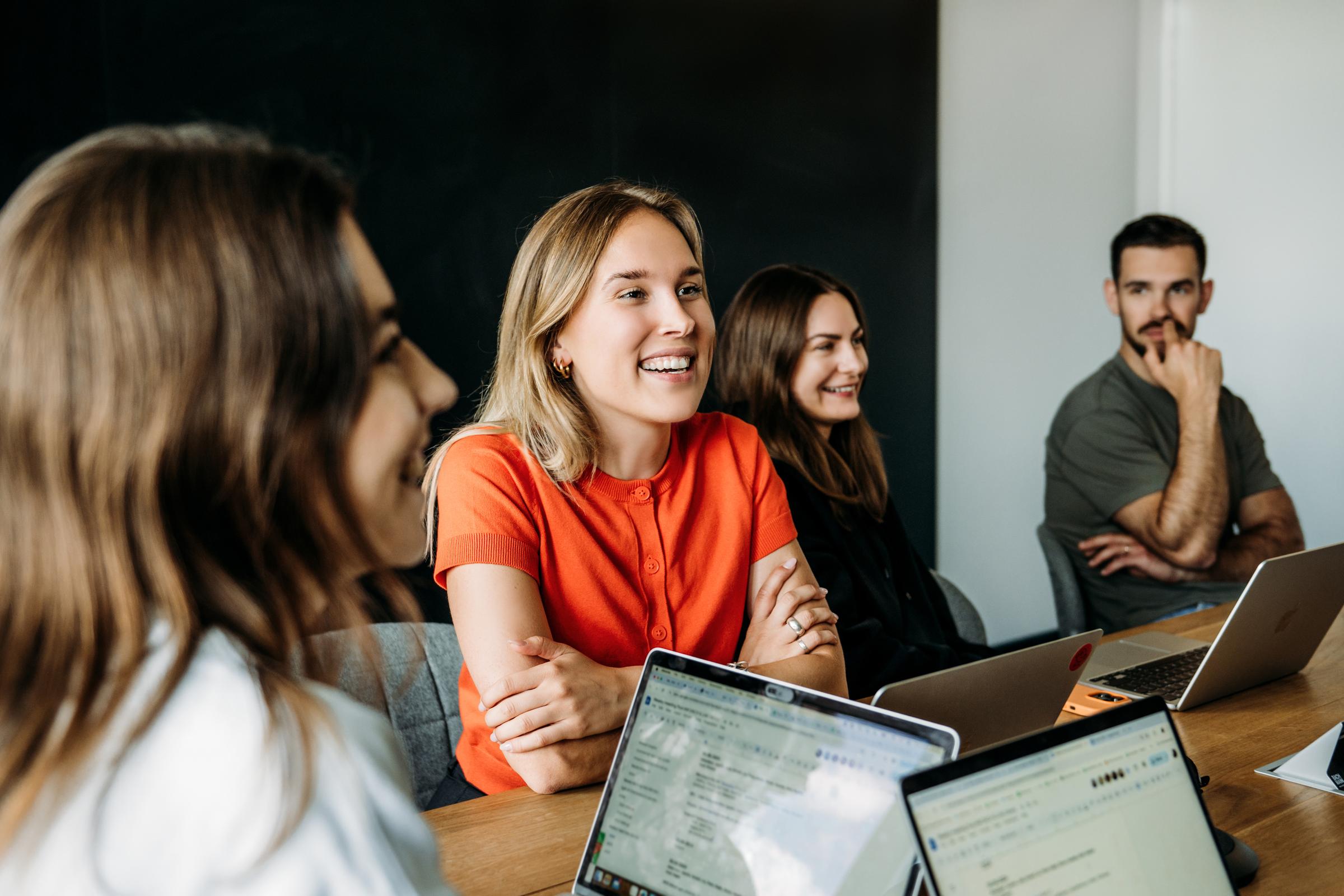 A women sitting behind her laptop and smiling accompanied by other colleagues 