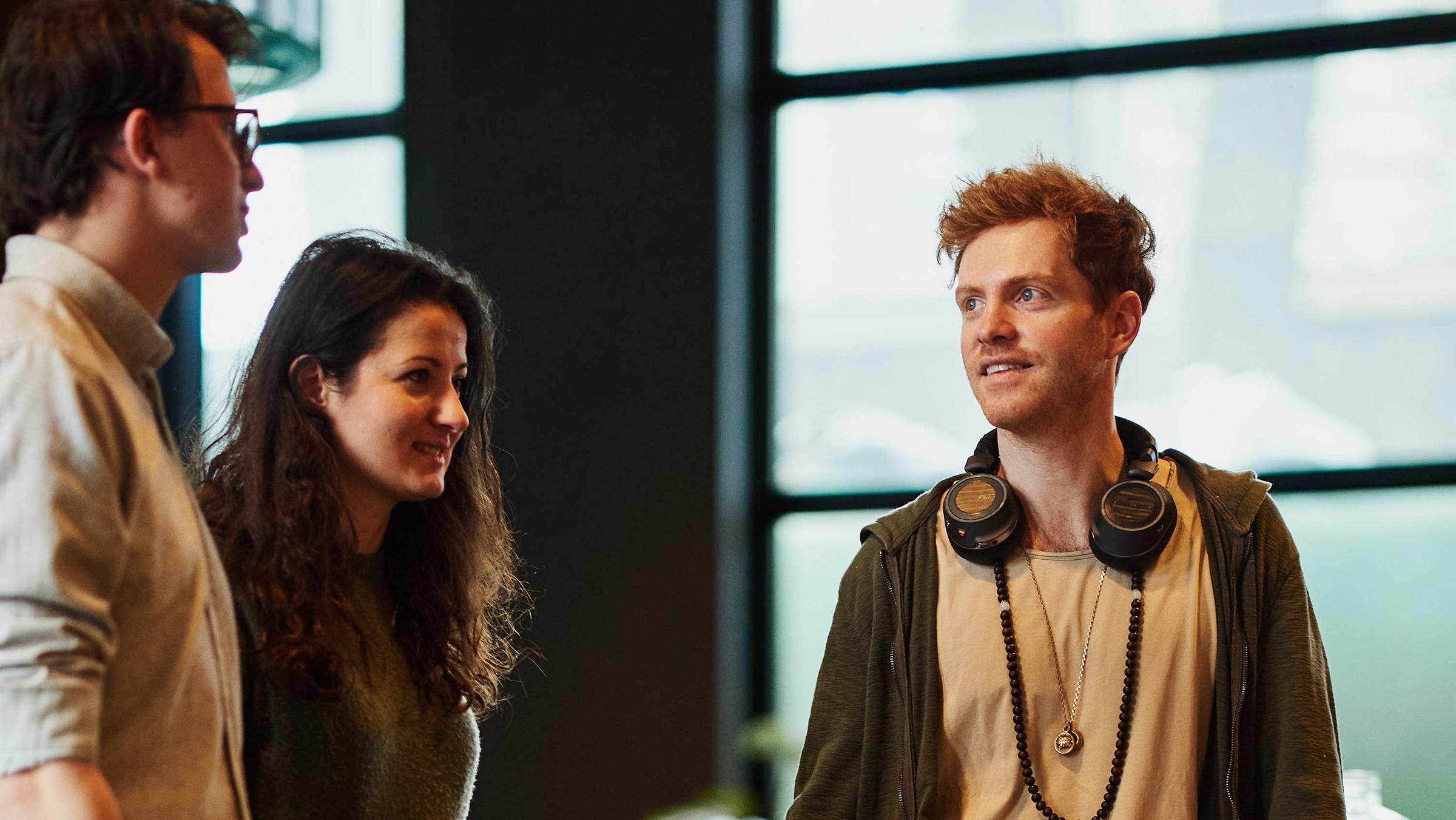 Three colleagues conversing in an office setting with large windows.