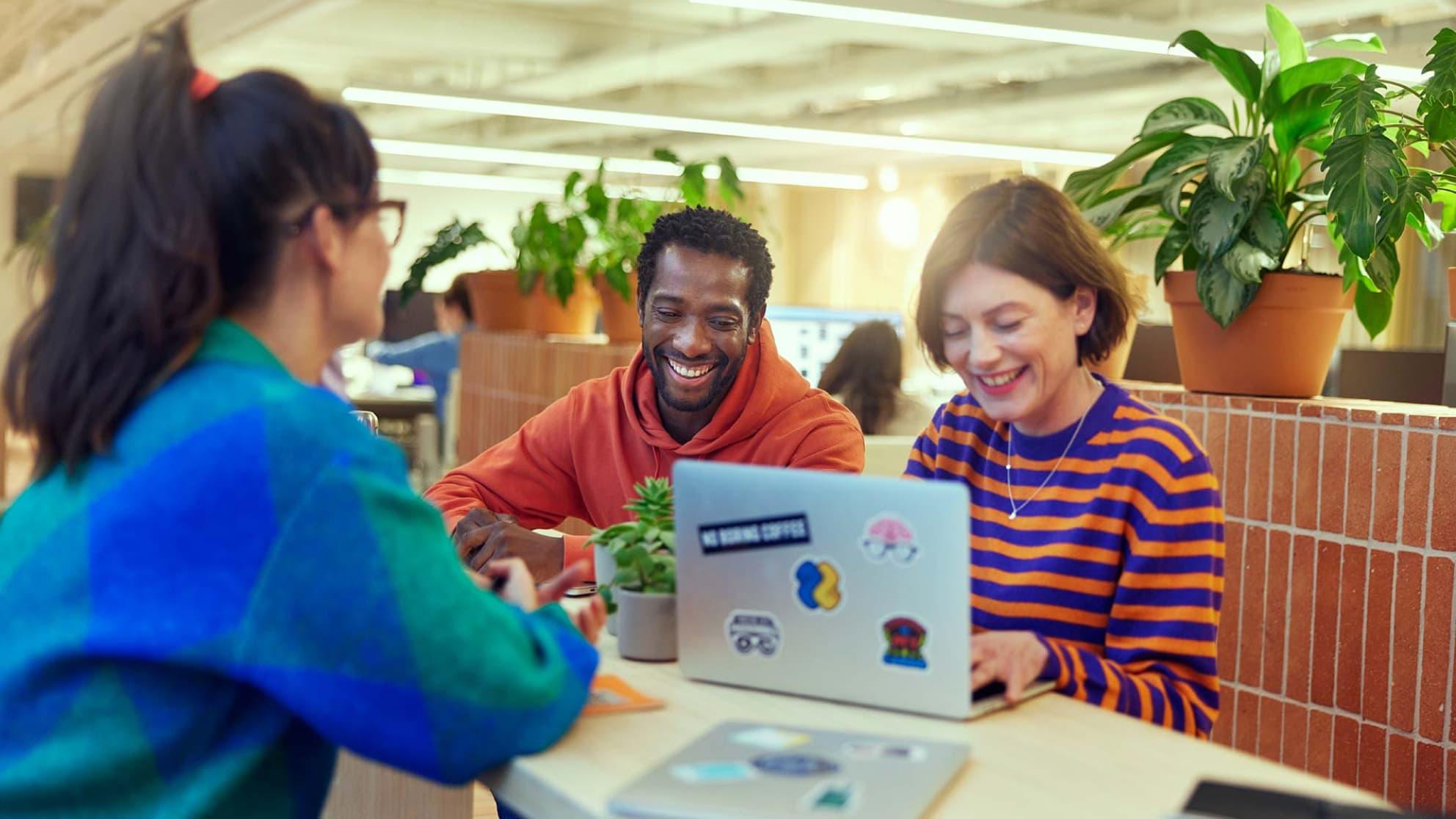 Três colegas sorrindo e trabalhando juntos com um laptop com adesivos Adyen.