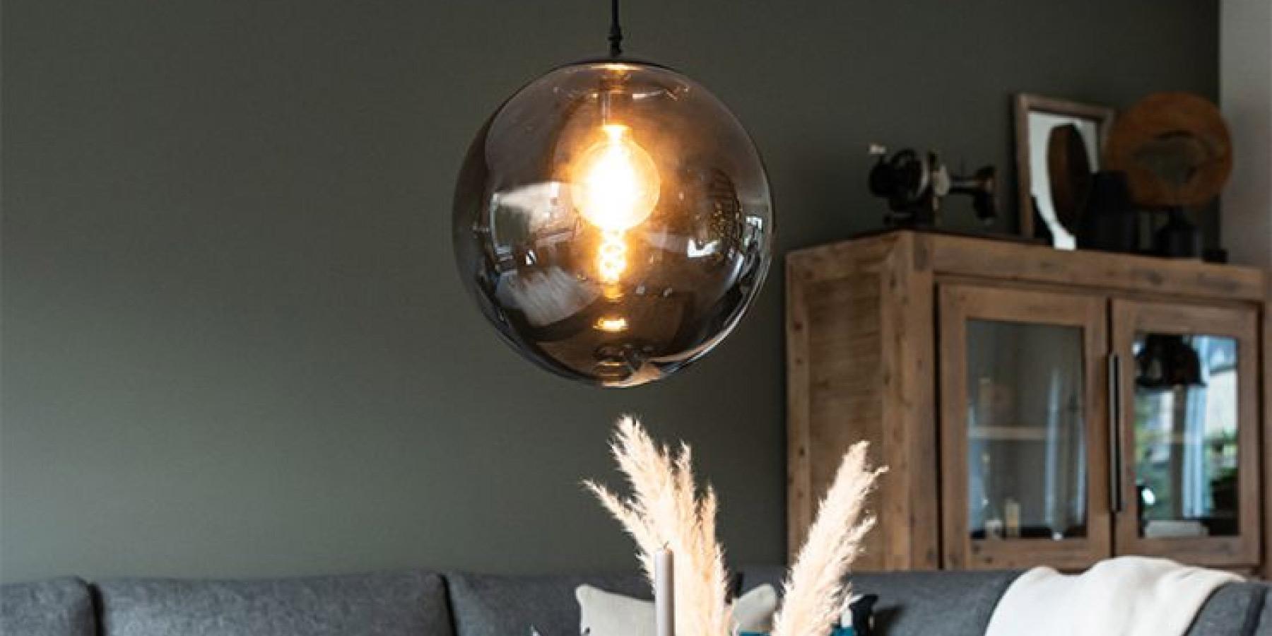 Moderne inrichting met een ronde glazen hanglamp boven een grijze bank, naast een houten kast.