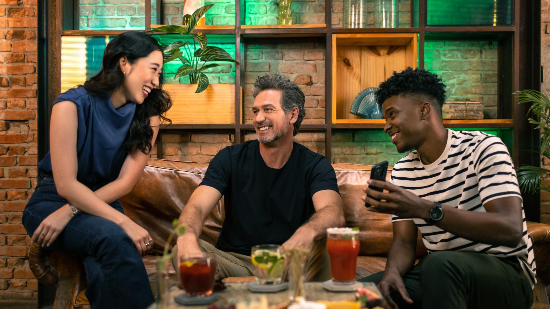 Três pessoas sorrindo e engajadas em uma conversa em um sofá com bebidas na mesa.