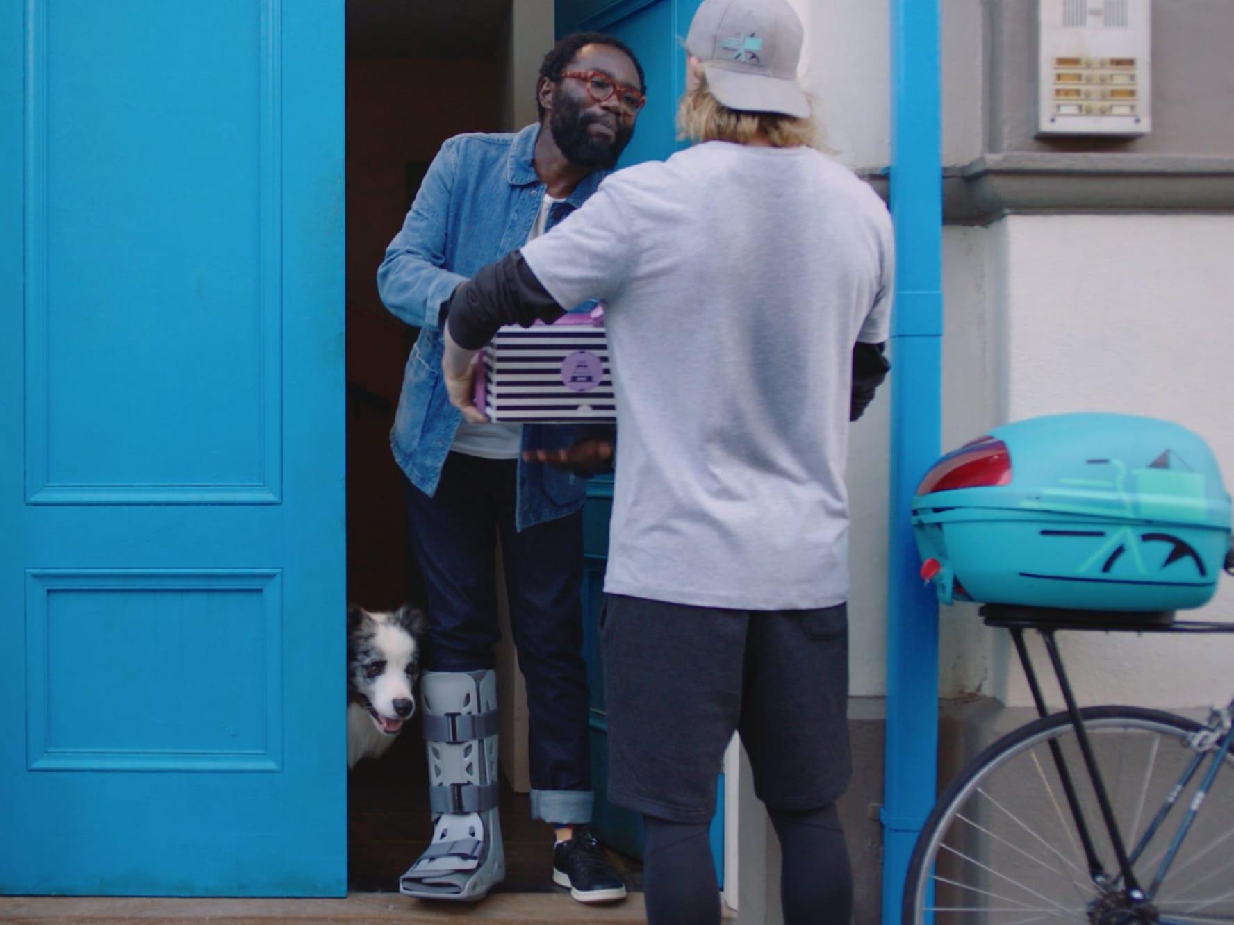 Persona entregando un paquete a un destinatario sonriente con un perro asomándose por la puerta.